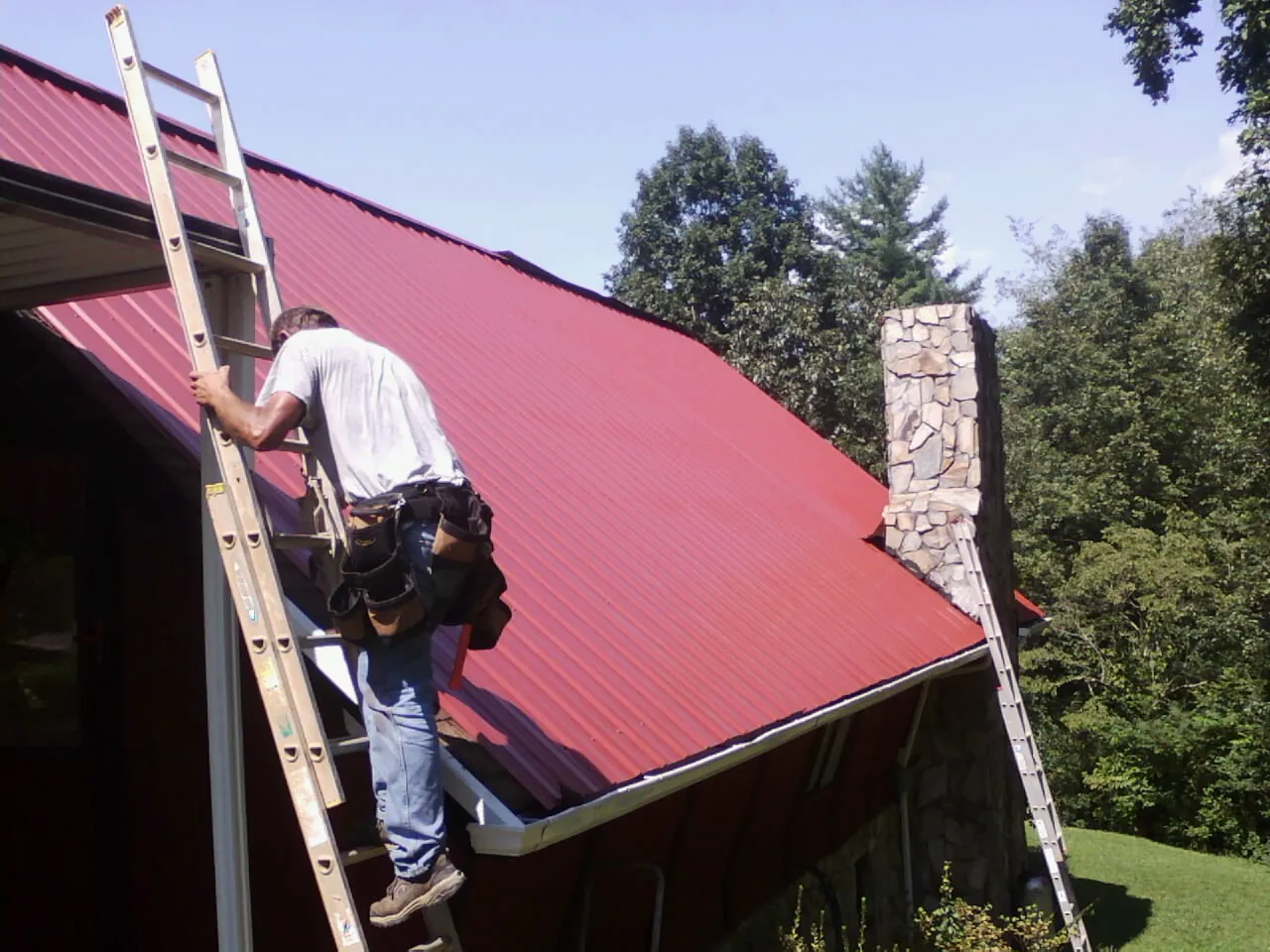 Roofer on a ladder beside a red metal roof and stone chimney — Soffit & Fascia Repair by Roofing Brothers in Cochranville